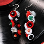 A pair of handmade dangle earrings featuring colorful felt-like rings and faux gemstones in red, black, white, green, and orange, displayed on a black vinyl record background. populonia