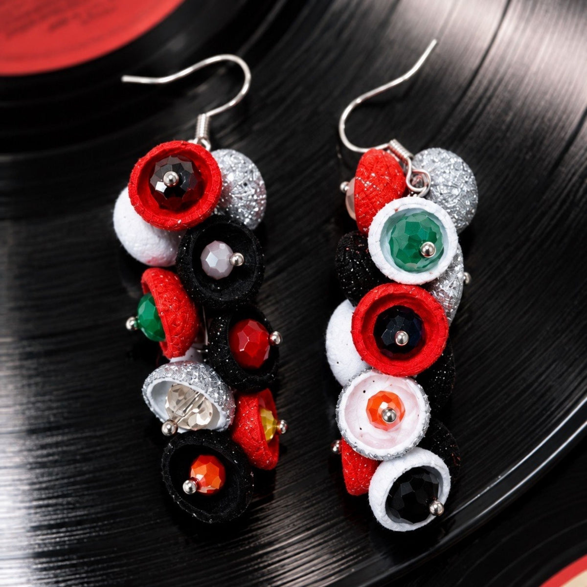 A pair of handmade dangle earrings featuring colorful felt-like rings and faux gemstones in red, black, white, green, and orange, displayed on a black vinyl record background. populonia