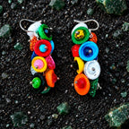 A pair of colorful dangling earrings made from stacked round and oval beads in bright rainbow colors, displayed on a dark textured surface with scattered green stones. populonia