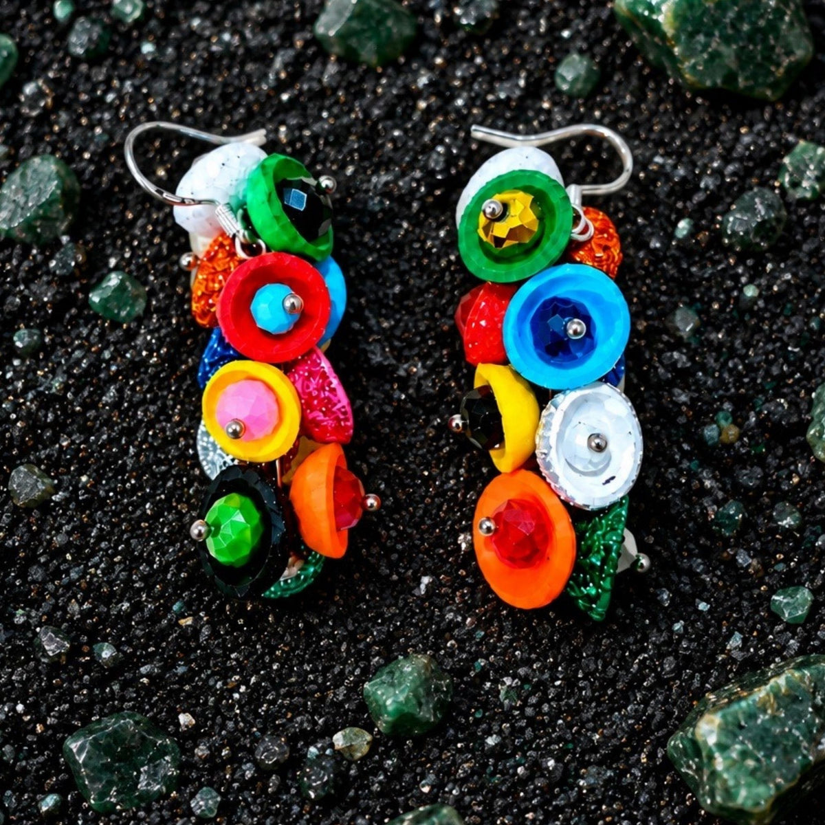 A pair of colorful dangling earrings made from stacked round and oval beads in bright rainbow colors, displayed on a dark textured surface with scattered green stones. populonia