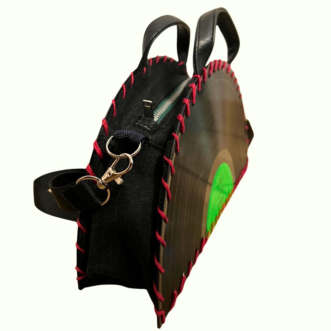 A black vinyl record shaped shoulder bag with red contrast stitching, a black canvas body, black leather handles, and a green label graphic on the front, shown on a plain white background.