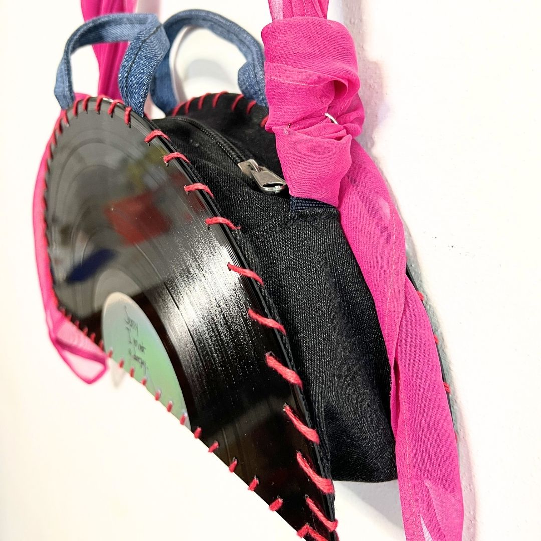 A handmade black fabric purse decorated with a vinyl record applique, pink sheer trim, and a bright pink ribbon handle, shown against a plain white background.