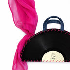 A novelty black vinyl record handbag with a denim blue handle, pink decorative stitching, and a bright pink sheer scarf tied to the handle, shown on a white background.