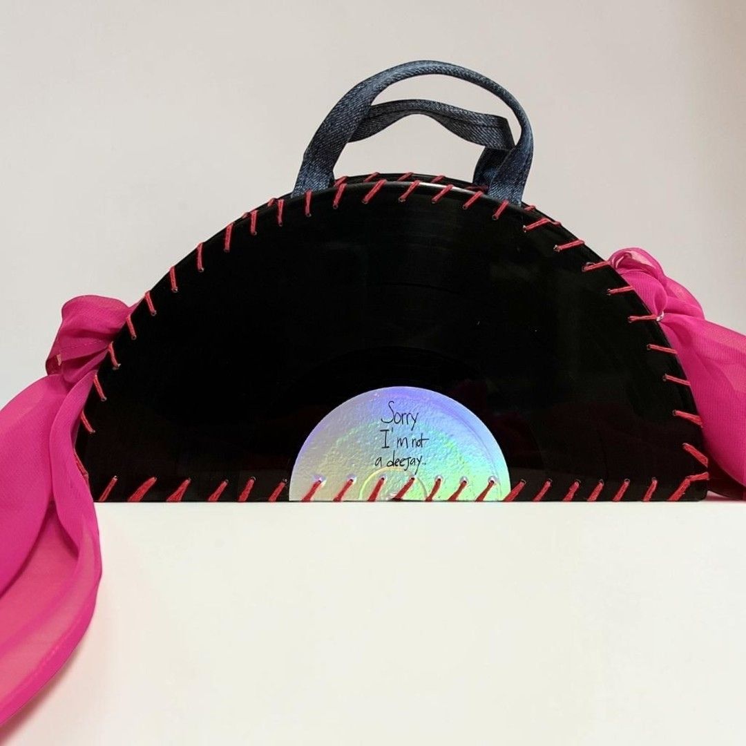 A handmade black vinyl record purse with red hand-stitched edges, a dark gray fabric handle, and bright pink fabric side flaps. A holographic sticker on the front reads 'Sorry I mad a scratch.