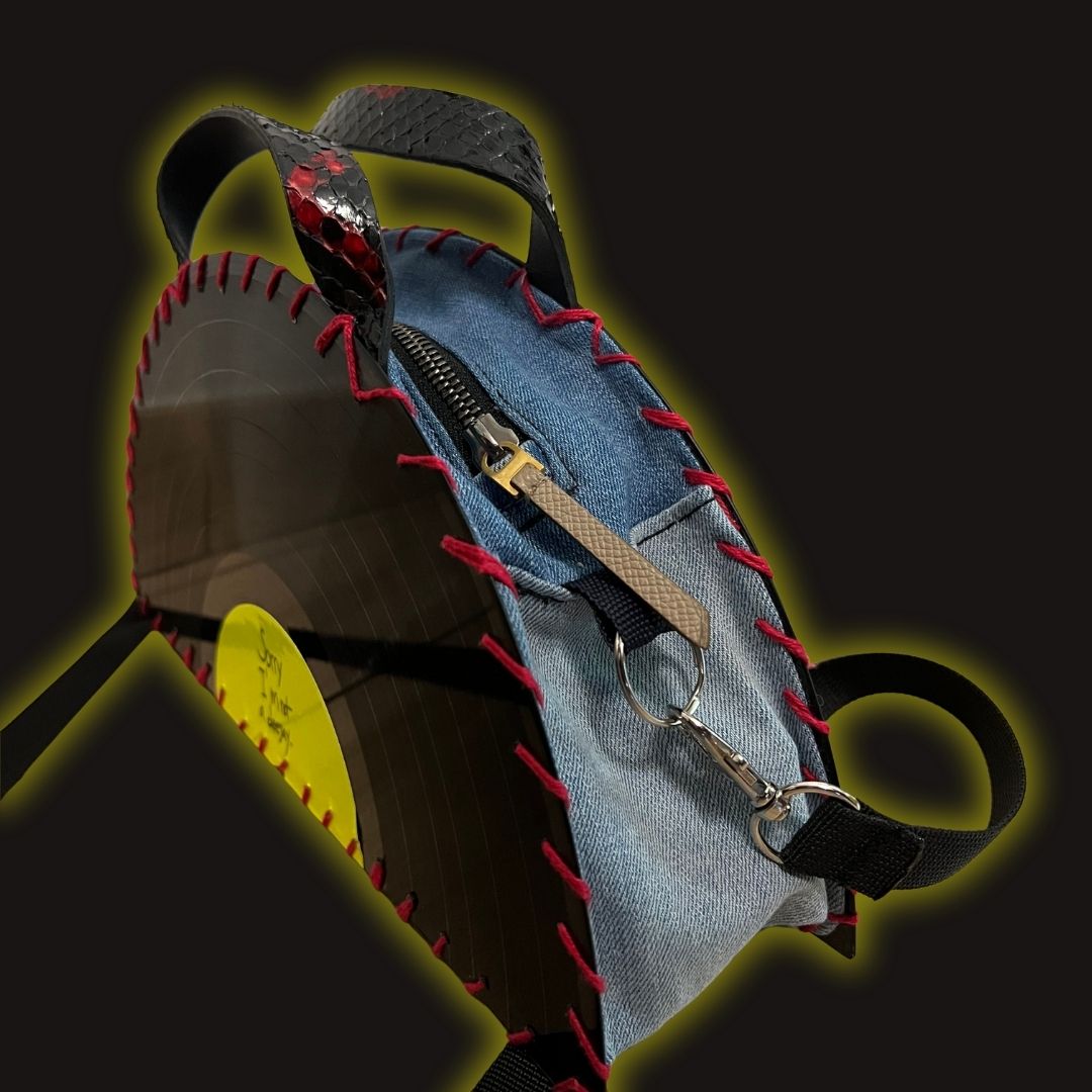 A circular denim and black vinyl record bag with red whipstitching, a zipper, and a keychain ring, shown on a dark background with a yellow glow effect.